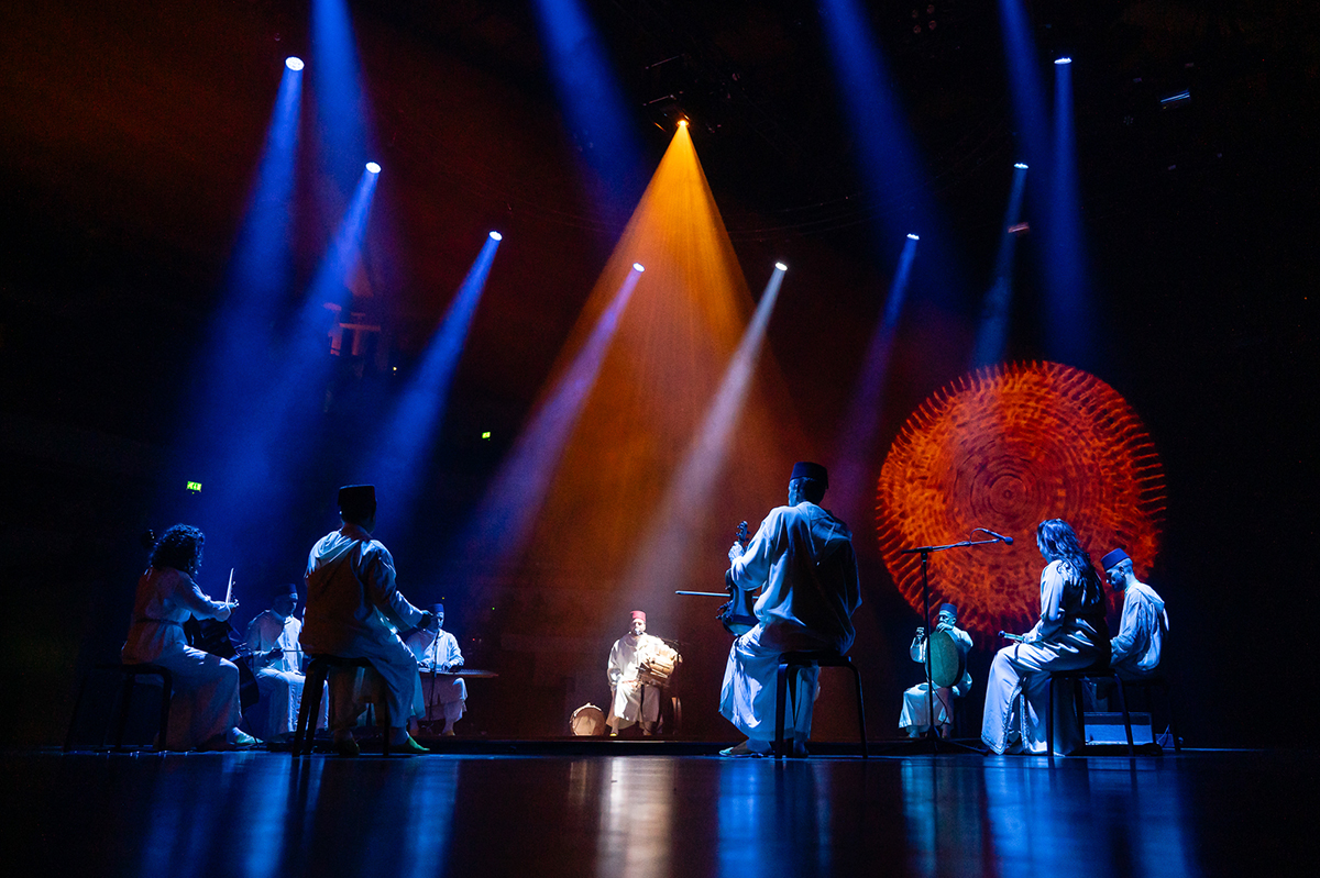 Several performers are seated on stage in blue light, while the singer is highlighted in white. Behind them, circular visuals are projected.